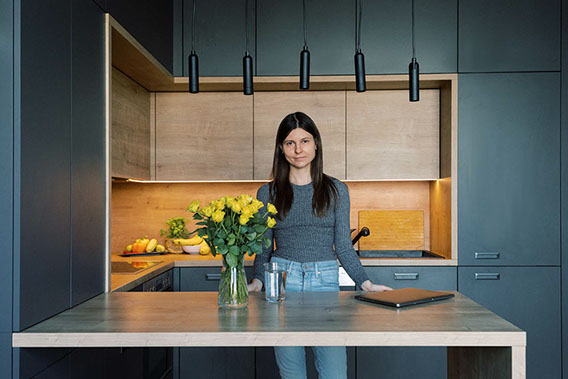 Woman standing in a modern kitchen with dark cabinetry and a wooden countertop, decorated with a vase of yellow flowers.