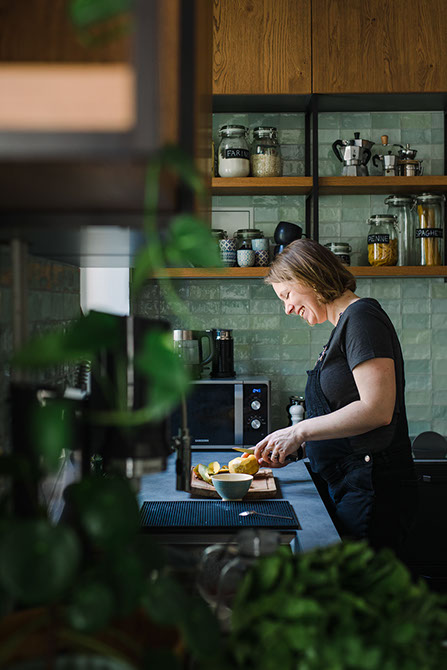 Woman cooking in a modern kitchen with green tiles, black cabinetry, and wooden shelving.