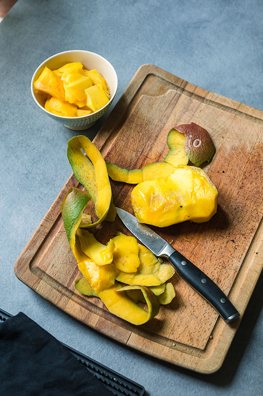 Freshly peeled mango on a wooden cutting board with a knife and a bowl of mango chunks.