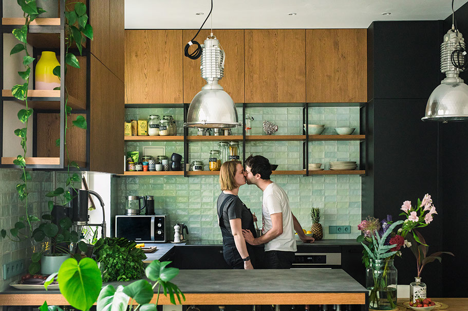 Couple sharing a kiss in a modern kitchen with green tile backsplash and open shelving.