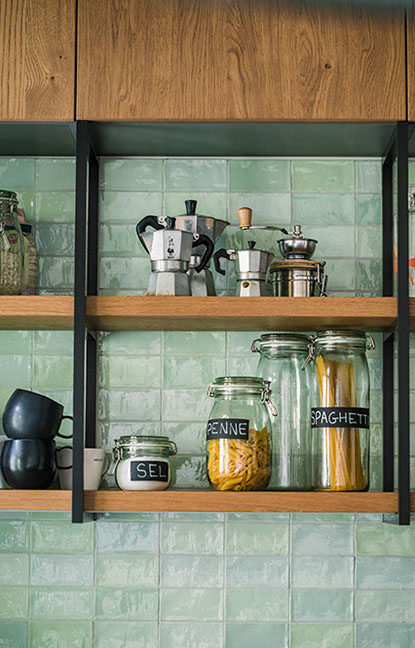 Open kitchen shelving with jars of pasta and coffee machines against a green tile backsplash.