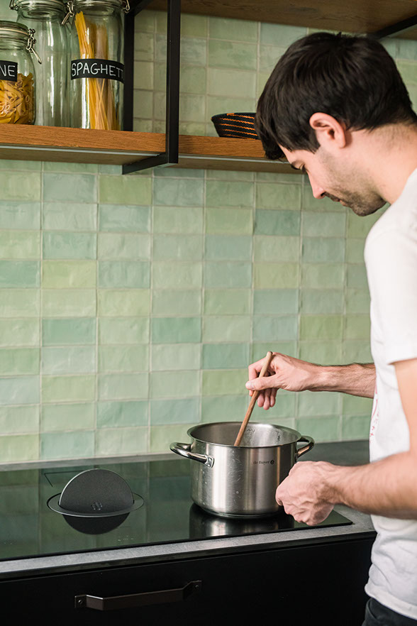 Man cooking in a modern kitchen with green tile backsplash and black cabinets.