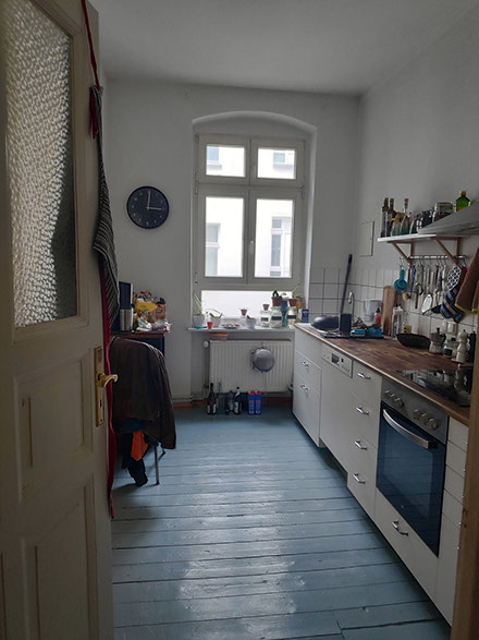 Small kitchen with blue wooden floors, white cabinetry, and a window overlooking the street.